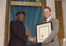 À cette occasion, Willie Brown, maire de San Francisco (à gauche) a présenté une proclamation à l’Église en saluant « ses efforts pour rendre la région de la baie un endroit meilleur pour tous, quels que soient leur origine, leur couleur, leurs croyances et leur milieu. »
